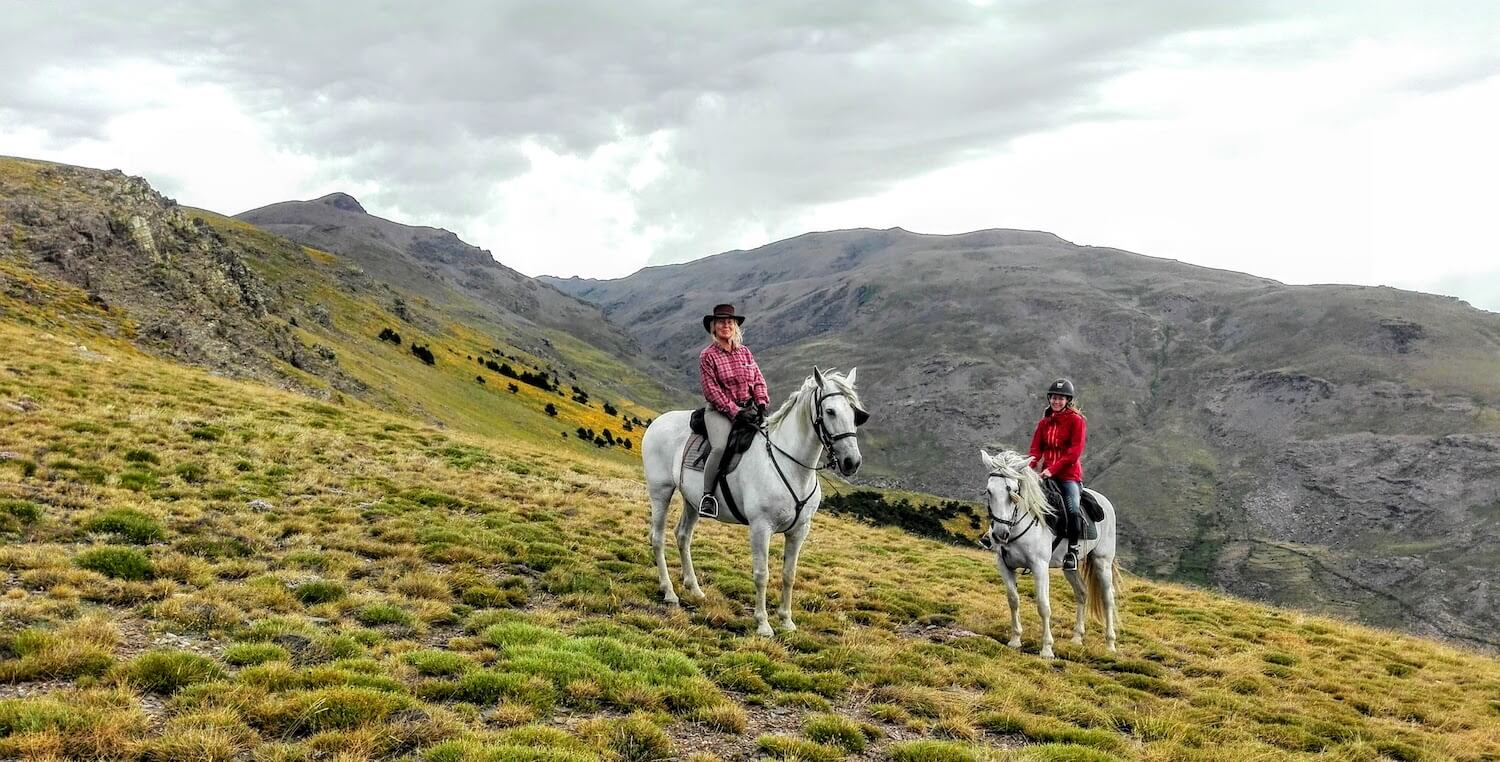 Two people on horseback riding towards a stunning mountain range.